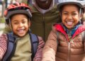 Amidst the hustle and bustle of the city, this heartwarming scene unfolds where a father, donning a casual cap and a warm smile, enjoys quality time with his kids during a bike ride. The children, each sporting protective helmets and vibrant winter jackets, clearly share their father's enthusiasm. Their beaming smiles and the close-knit atmosphere suggest a strong bond and the value of family activities. It's a beautiful image that reflects parenting at its best – engaging, safe, and full of joy.