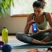 In the tranquil setting of her home gym, a woman takes a moment to sit cross-legged on her yoga mat. Surrounded by fitness equipment like dumbbells, a kettlebell, and a water bottle, she focuses intently on her smartphone. She is likely engaged in selecting a new workout routine or setting fitness goals using a specialized fitness app. The atmosphere suggests a dedication to personal health and wellness, combining technology with traditional exercise tools.