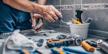 In this photo, a handyman is captured in the middle of a meticulous home improvement task, demonstrating precision and skill. Surrounded by an assortment of tools ranging from wrenches to screwdrivers, his focus is on repairing or installing components essential for the project. The cluttered yet organized workspace reflects the complexity and nature of his work, emphasizing a professional who is well-versed in managing multiple aspects of construction and maintenance efficiently.