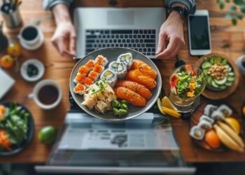 In the midst of a bustling workday, a professional sits at a cluttered desk adorned with various digital devices and personal items, indulging in a nutritious and visually appealing meal. The lunch features an assortment of sushi, including nigiri and rolls, accompanied by a fresh salad and a selection of fruits, showcasing a balance between indulgence and health. This setting illustrates the modern blend of work and wellbeing, where productivity meets self-care.
