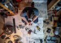 This image captures a top-down view of an inventive individual deeply engrossed in assembling or repairing electronic devices in a workshop. The workspace is strewn with various tools, electronic components, and schematics, reflecting a scene of creative chaos. The presence of soldering equipment, drone parts, and multiple projects in mid-construction suggests a blend of engineering prowess and a passion for technology. This vibrant depiction highlights the intense focus and intricate details involved in the technical crafting process.