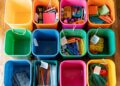 A top-down view of vibrantly colored storage bins neatly positioned on a polished wooden floor. Each bin is filled with an assortment of school supplies, showcasing a methodical approach to organization. From art supplies to notebooks and writing instruments, the array of items is meticulously sorted by category, making it easy to find what's needed. The image exudes a sense of order and preparedness, ideal for a classroom or a home study area.