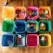 A top-down view of vibrantly colored storage bins neatly positioned on a polished wooden floor. Each bin is filled with an assortment of school supplies, showcasing a methodical approach to organization. From art supplies to notebooks and writing instruments, the array of items is meticulously sorted by category, making it easy to find what's needed. The image exudes a sense of order and preparedness, ideal for a classroom or a home study area.