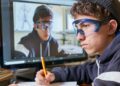In this image, a young male student is seen deeply absorbed in his studies. He is seated at a desk with a pencil in hand, working on his notes while looking intently at a computer screen displaying the image of another person wearing protective goggles. This suggests he might be involved in an online educational workshop or a science class. His expression, coupled with the educational setting, highlights the importance of digital learning tools in enhancing student engagement and learning outcomes.
