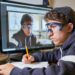 In this image, a young male student is seen deeply absorbed in his studies. He is seated at a desk with a pencil in hand, working on his notes while looking intently at a computer screen displaying the image of another person wearing protective goggles. This suggests he might be involved in an online educational workshop or a science class. His expression, coupled with the educational setting, highlights the importance of digital learning tools in enhancing student engagement and learning outcomes.